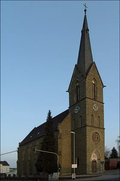 Kath. Kirche St. Antonius Eremita in 65552 Limburg an der Lahn - Eschhofen Eschhofen besaß bis zum 1895 erfolgten Abbruch eine spätmittelalterlichen Antoniuskapelle im älteren Ortskern. Den Neubauplan lieferte 1888 Diözesanbaumeister Jakob Fachinger aus Limburg, der damit einen anspruchsvolleren Entwurf Max Meckels verdrängte. Aber auch der Entwurf Fachingers wurde nur mit einem niedrigeren Turm und vermutlich kürzerem Chor ausgeführt. Die Weihe erfolgte 1891. Die neugotische Saalkirche aus unverputztem grünem Schalstein zeigt einen mittigen Fassadenturm mit Maßwerkrosette an der Westseite und einen kleinen, eingezogenen 5/8-Chor. Das schlichte, harmonisch proportionierte Äußere mit gestuften Strebepfeilern und großen Maßwerkfenstern setzt einen deutlichen Akzent in der Abfolge der Häuserzeile der fast schnurgeraden Limburger Straße. Im Tympanonfeld des spitzbogigen Turmportals Wandgemälde mit einem thronenden Christus in der Mandorla, der von Maria und Johannes flankiert wird. Das Innere besitzt durchgängig querrechteckige, tiefgefaltete Kreuzrippengewölbe auf zierlichen Konsolen ohne Hervorhebung der Gurtrippen. Die ursprüngliche Ausmalung von 1892 ist seit dem Weißanstrich von 1953 verschwunden. Bis auf die Orgelempore wurde die gesamte Ausstattung erneuert einschließlich der Chorfenster. Zur Sachgesamtheit gehören die Umfassungsmäuerchen mit Gittern aus gereihten Spitzbogenformen. Seit Errichtung der Pfarrei „Heilige Katharina Kasper Limburger Land (Sitz: Limburg a. d. Lahn)“ zum 1. Januar 2023 ist die Kirche nicht mehr im Rang einer Pfarrkirche. (Denkmalpflege-Hessen.de)