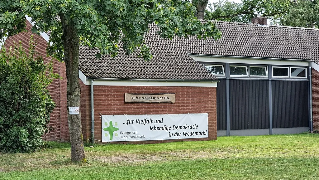 Die Auferstehungskirche Elze in der Gemeine Wedemark, Region Hannover, hier mit dem am Kirchenbau aufgespannten Banner „... für Vielfalt und lebendige Demokratie in der Wedemark!“ links davor das Logo und das dazugehörige Motto „Evangelisch in der Wedemark“ ...