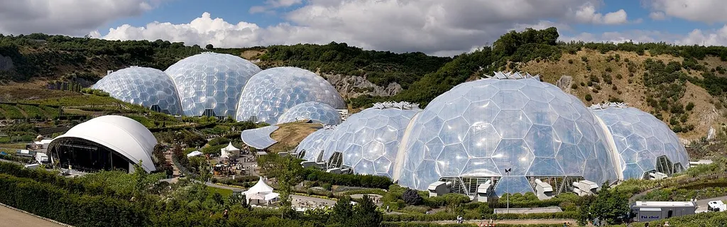 Panoramic view of the geodesic dome structures of the Eden Project. The Eden Project is a large-scale environmental complex near St Austell, Cornwall, England, United Kingdom. The project was conceived by Tim Smit and has quickly become one of the most popular visitor attractions in the United Kingdom. The complex includes two giant, translucent domes made of ETFE cushions, each emulating a natural biome, that house plant species from around the world. The first emulates a tropical environment, the other a warm temperate, mediterranean environment. The project took 2½ years to construct and opened to the public in March 2001.