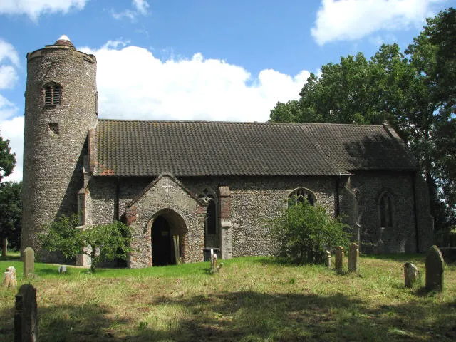 All Saints Church - view across churchyard. The round tower church of All Saints, restored in Victorian times, has a number of interesting medieval survivals such as the rood stairway > 629913; an interesting and unique 15th century baptismal font > 721790 - 721801, the panels of which were recoloured in the 1930s, depicting saints on panels and stem and a wall painting depicting the narrative of St Christopher, believed by some to be the finest in all England > 629910. A 13th century coffin slab, now serving as a bench, is inserted into the south wall > 721789. The church is kept locked but a key is available. For more information see: http://www.norfolkchurches.co.uk/hemblington/hemblington.htm
